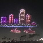 An aerial drone light show depicts a glowing cityscape with a two-boat formation below it, showcasing the spectacle of the St. Pete Fall Festival.