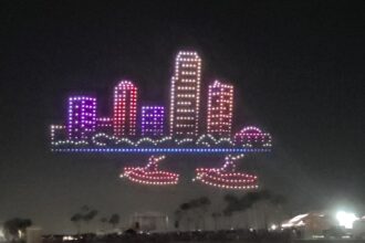 An aerial drone light show depicts a glowing cityscape with a two-boat formation below it, showcasing the spectacle of the St. Pete Fall Festival.