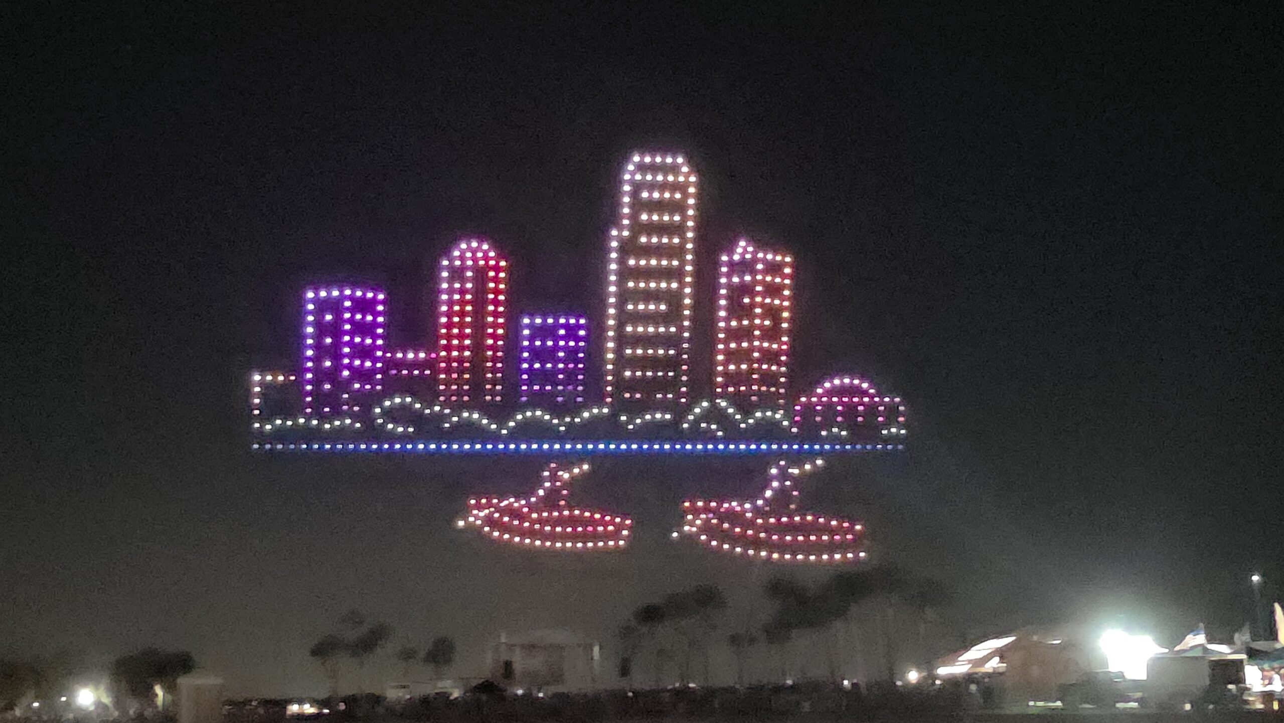 An aerial drone light show depicts a glowing cityscape with a two-boat formation below it, showcasing the spectacle of the St. Pete Fall Festival.