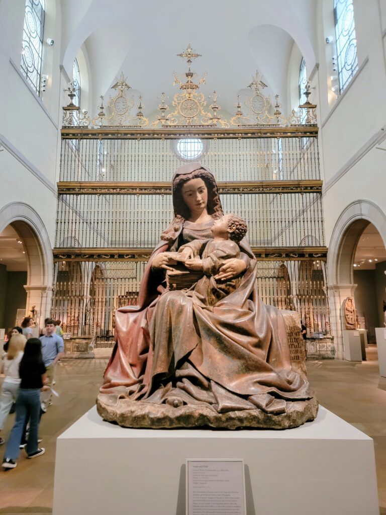 Sculpture of a seated woman holding a child in a museum gallery with ornate ironwork and arched windows—highlighting religious art and serene atmosphere.
