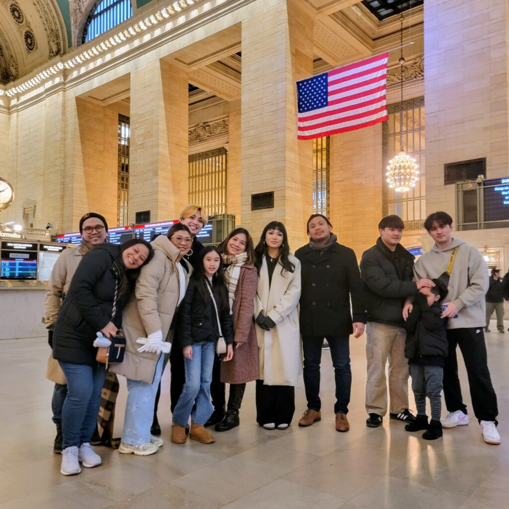 Group of eleven people posing inside Grand Central Terminal with ornate chandeliers, American flag, and train boards—celebrating connection in a historic NYC landmark.