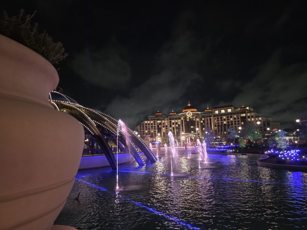 A nighttime view of a grand hotel at Universal’s Epic Universe, with a vibrant water fountain display in front. The fountains glow in purple and blue hues, while the multi-story building behind is warmly lit, showcasing ornate architecture and a luxurious ambiance.