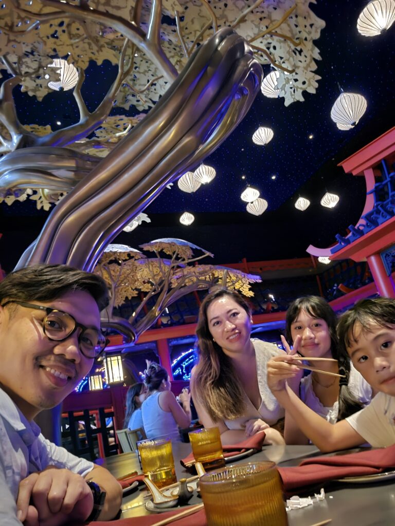 A family of four sitting at a dining table in a restaurant with a whimsical decor featuring a large artistic tree and decorative lanterns, smiling and enjoying their meal.