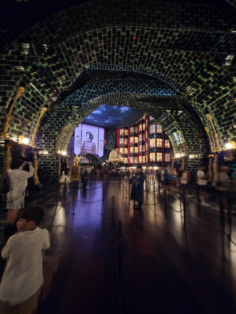 An interior view of a darkly lit venue featuring a green tiled archway, with visitors walking and taking photos, and colorful screens in the background.