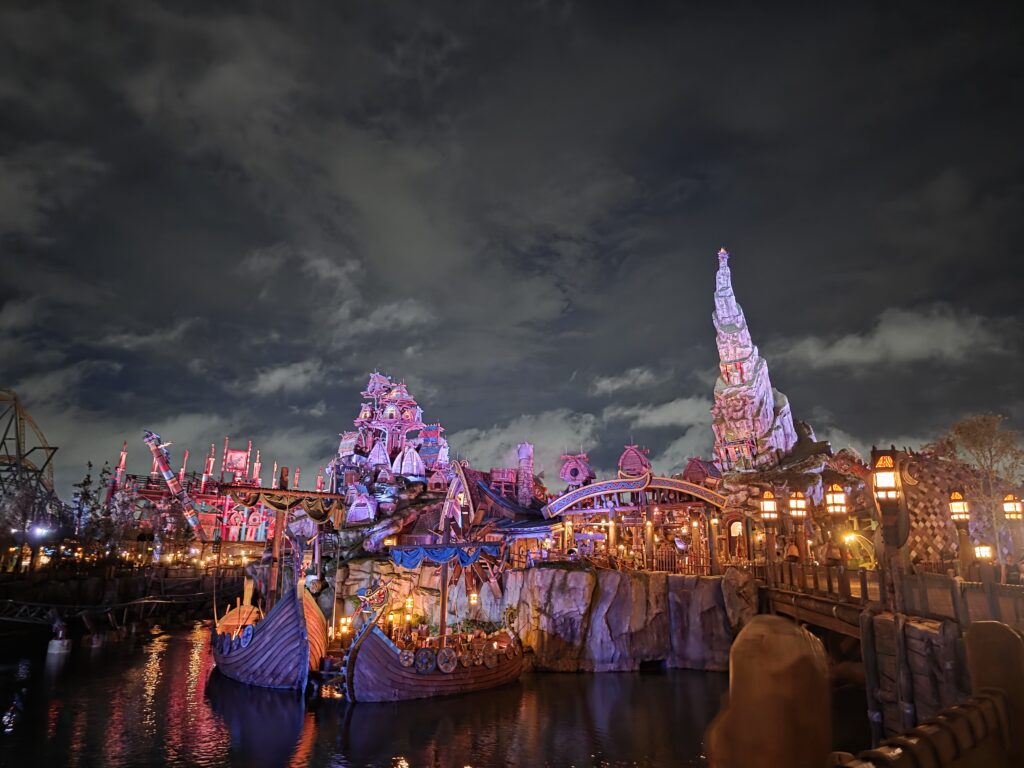 A nighttime view of a fantastical theme park with illuminated buildings and a large rocky structure, featuring Viking ships on a waterway, surrounded by colorful lights and dramatic clouds.