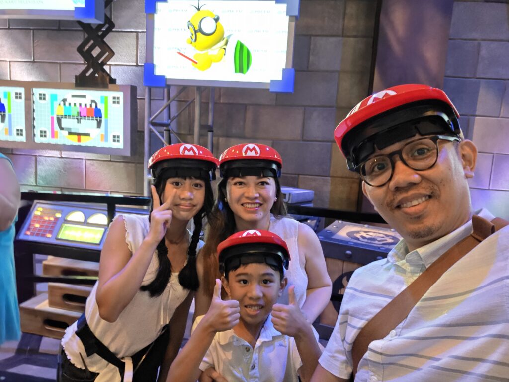 A family of four wearing Mario-themed AR headsets, smiling and posing for a selfie in a colorful setting with gaming screens in the background.
