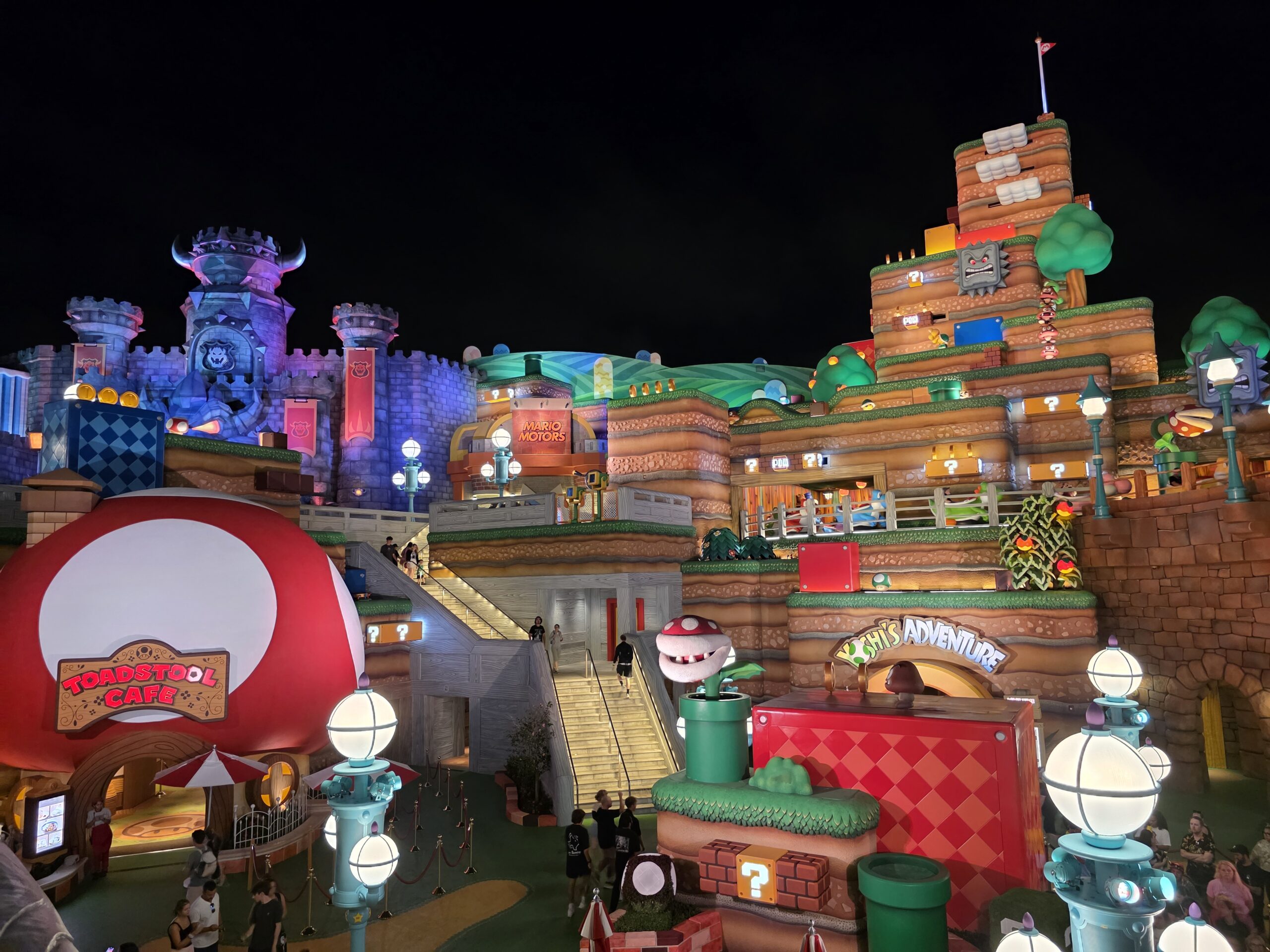 Nighttime view of Super Nintendo World at Universal's Epic Universe, featuring colorful buildings, Power-Up items, and the Toadstool Cafe, with visitors exploring the vibrant area.