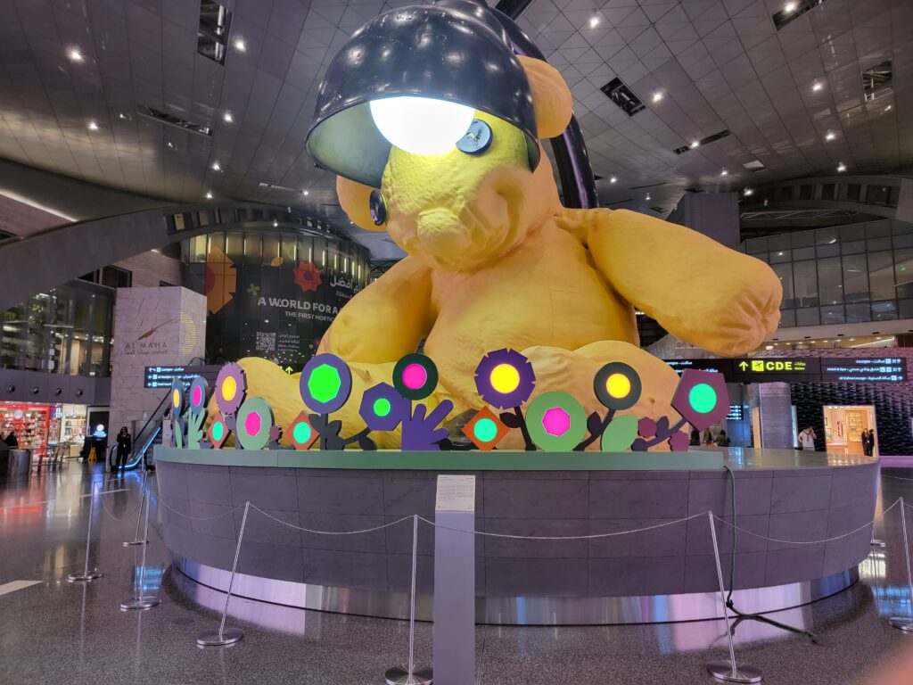 Giant yellow teddy bear sculpture with a desk lamp embedded in its head, located in Hamad International Airport—an iconic art piece representing the best of Qatar’s cultural flair