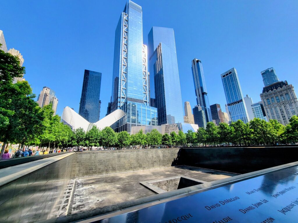 9/11 Memorial reflecting pool with engraved names, One World Trade Center, and the Oculus in the background—commemorating lives lost and NYC’s strength.